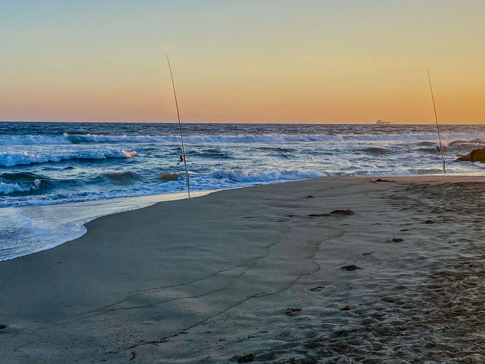 Surf rods planted in wet sand at sunset, waves rolling in behind them