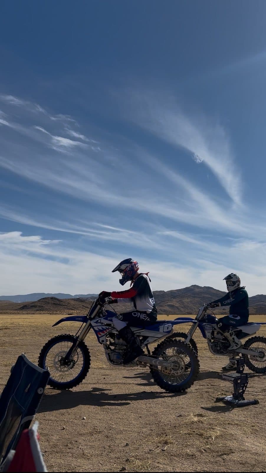 Two helmeted dirt bike riders waiting on an open desert flat under a wide streaked sky